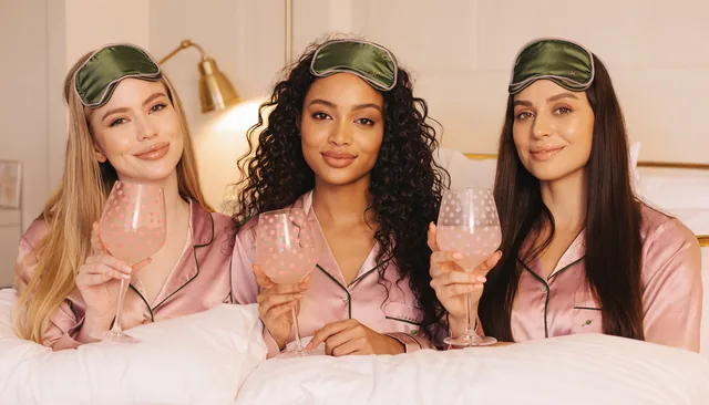 Three friends in matching blush satin pyjamas piped in olive, with cream shell buttons and an olive on a pick embroidered on the pocket, their olive eye masks piped in blush pushed up, leaning on white pillows and smiling, each holding a polka-dot painted glass of pink 0.0%