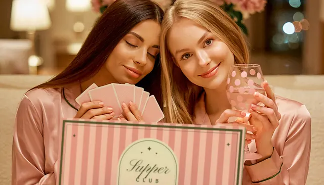 Two friends in blush satin pyjamas, cheek to cheek: one with her eyes closed, holding her cards to her heart, the other smiling over a polka-dot painted glass of pink 0.0%, the striped Supper Club lid open in front of them
