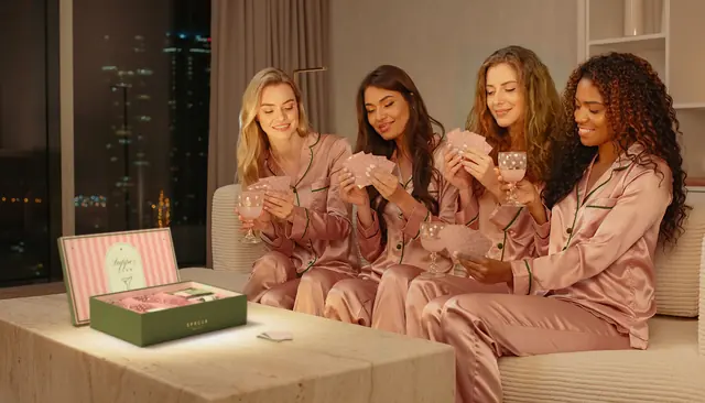 Four friends in matching blush satin pyjamas piped in olive on a cream sofa at night, the city lights in the window, holding their cards and polka-dot painted glasses of pink 0.0%, the open SPRCLB box on the marble coffee table